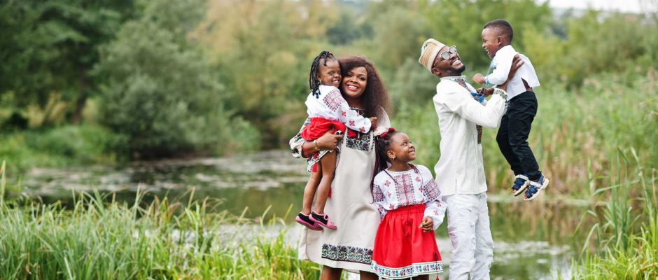 A Black family stands together on open land with greenery and distant trees in the background. The family faces the camera confidently, symbolizing opportunity, stability, and long-term settlement. The scene reflects land use, community, and future planning rather than formal ownership.