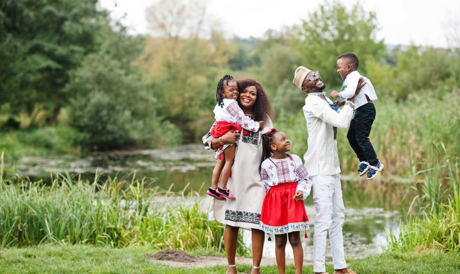 A Black family stands together on open land with greenery and distant trees in the background. The family faces the camera confidently, symbolizing opportunity, stability, and long-term settlement. The scene reflects land use, community, and future planning rather than formal ownership.