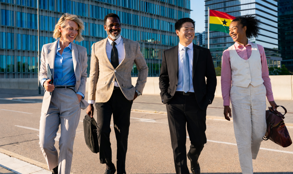 Four diverse international business professionals walking outside a modern office building in Ghana, with the Ghana flag mounted on a pole in the background, representing foreign investment and work opportunities in Ghana.