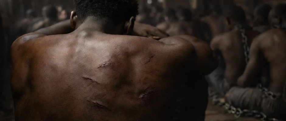 Enslaved African man with scars on his back aboard a slave ship, with chained captives blurred in the background beneath deck