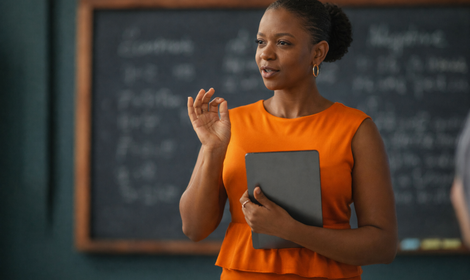 Ghanaian woman teacher in classroom holding tablet wearing orange dress