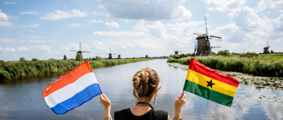 Woman holding Ghana flag and Netherlands flag beside canal with Dutch windmills in the background
