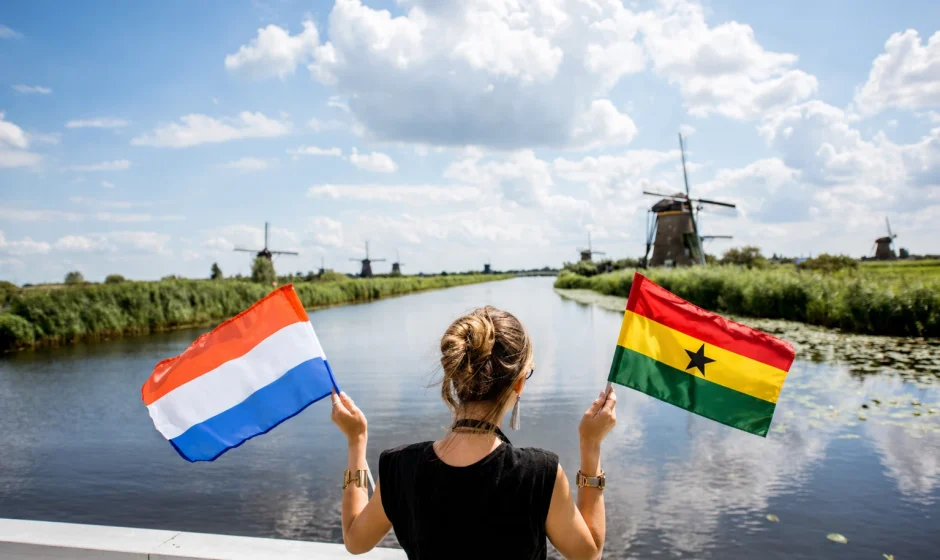 Woman holding Ghana flag and Netherlands flag beside canal with Dutch windmills in the background