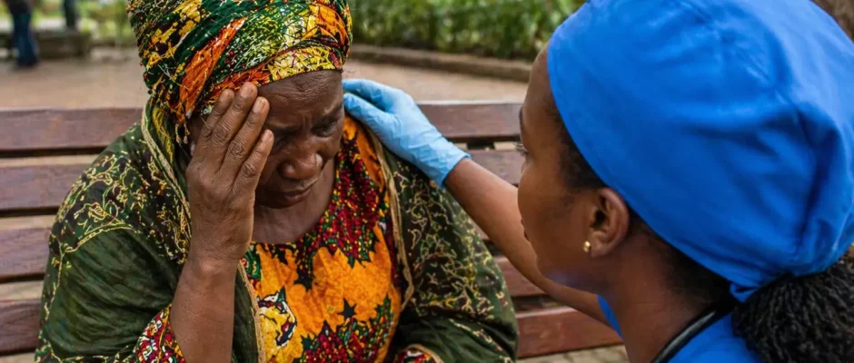 Elderly Ghanaian woman holding her head while a Ghanaian nurse assists her on a public bench in Kumasi, representing stroke deaths in Ghana and urgent medical response