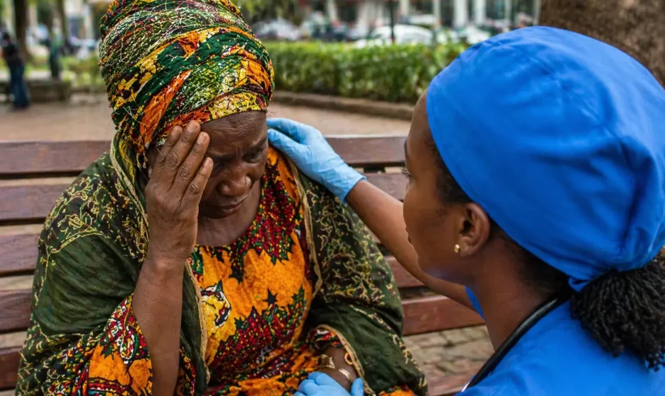 Elderly Ghanaian woman holding her head while a Ghanaian nurse assists her on a public bench in Kumasi, representing stroke deaths in Ghana and urgent medical response